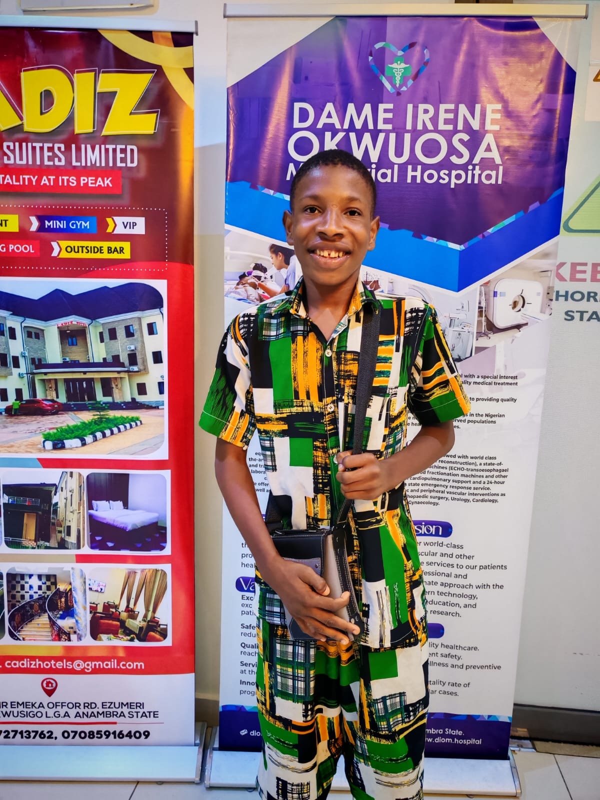A smiling young patient in a patterned green, yellow, and white outfit standing in front of hospital banners