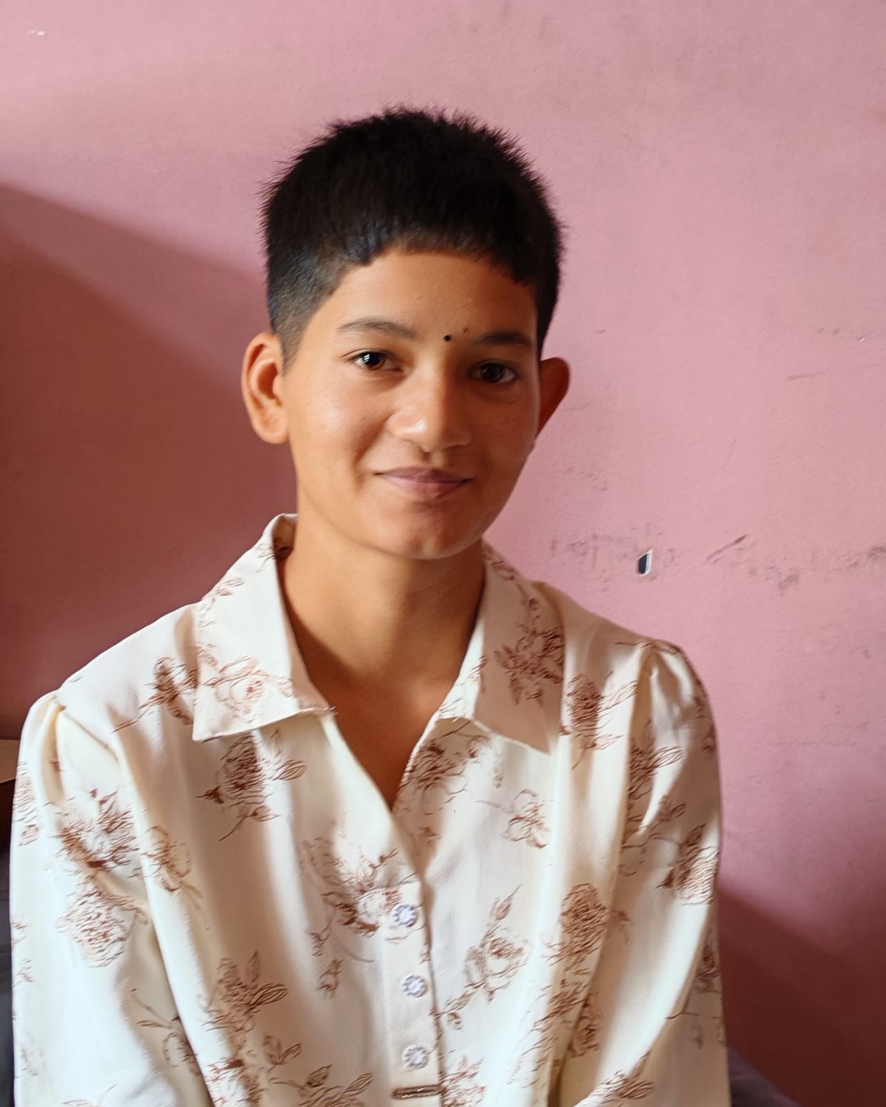 A young person in a cream floral blouse standing against a pink wall